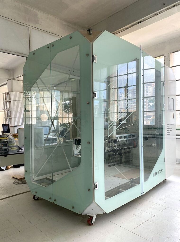 Big Voxel 2.0 large-format 3D printer: a tall enclosed fabrication cabinet with pale green glass panels on a steel frame, standing on castors in a workshop, with the print bed and gantry visible through the doors.